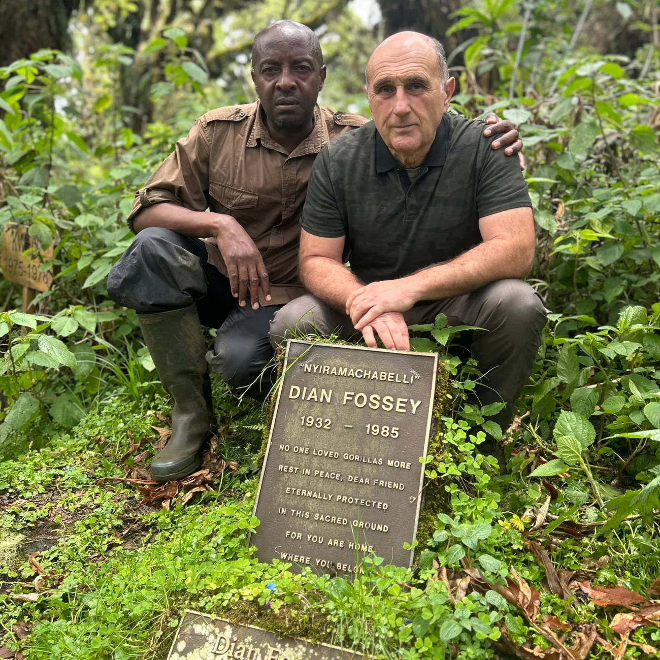 Fabrice Martinez et Jean Bosco Bizumuremyi accroupis derrière la plaque funéraire de Dian Fossey, dans la végétation